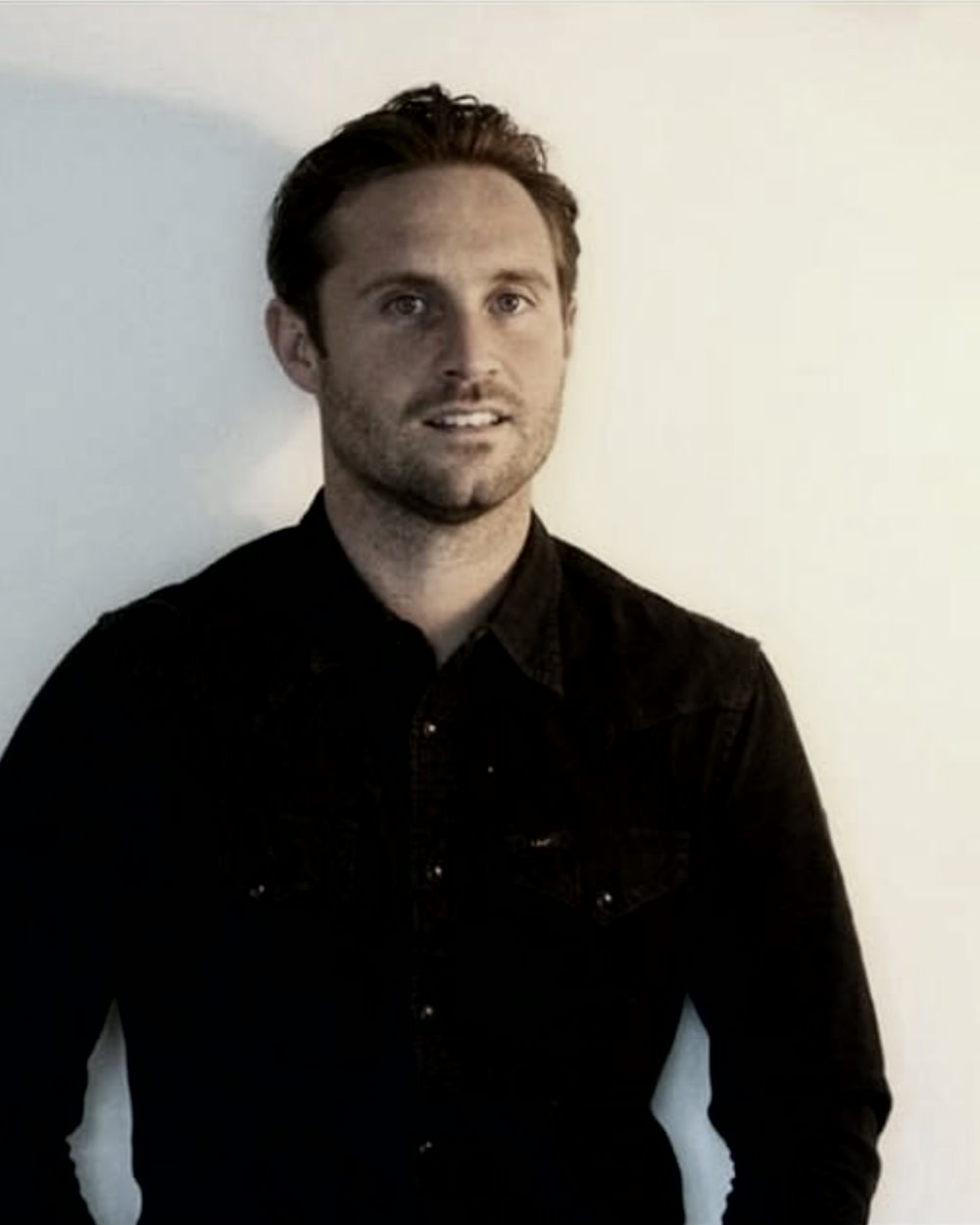 Mark, in a dark shirt against a pale wall, looking past the camera.